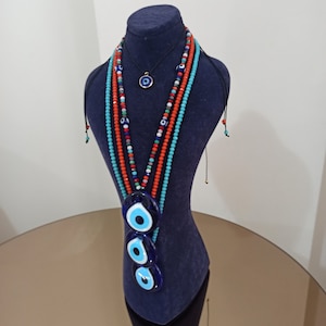 May include: A blue velvet display stand with three layered evil eye pendants on a beaded necklace. The necklace is made of turquoise, red, and white beads.