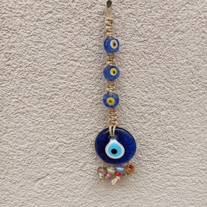 May include: A blue evil eye charm with a white center and yellow pupil, hanging from a string of beads and a knotted rope. The charm is attached to a white wall.