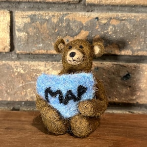 May include: A handmade brown teddy bear figurine holding a blue heart-shaped sign with the word "MAP" in black letters. The bear is made of a textured material and sits on a wooden surface.
