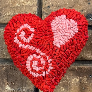 May include: A handcrafted, red heart-shaped decoration with a textured, looped design. The heart features two pink spiral patterns and a pink heart shape. The decoration is mounted on a silver metal rod, perfect for Valentine's Day or other special occasions.