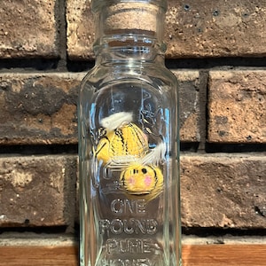 May include: A clear glass jar with a cork lid and a label that reads "One Pound Pure Honey". The jar is filled with honey and has a bee design on the front.