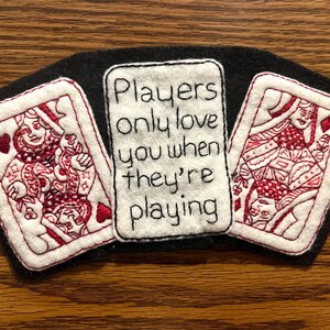 Op de afbeelding: Een zwart vilt patch met drie geborduurde elementen. Twee speelkaarten met de Hartenkoningin flankeren een centrale rechthoek met de tekst "Players only love you when they're playing" in zwart.
