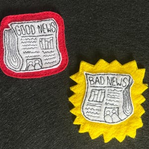 Pode incluir: Dois emblemas de feltro com desenhos de jornais bordados. Um emblema é um quadrado vermelho com cantos arredondados contendo um jornal branco com a inscrição GOOD NEWS. O outro é uma forma de estrela amarela com um jornal branco BAD NEWS.