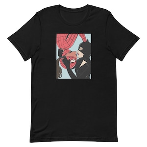 Puede incluir: Camiseta negra con una ilustración estilo cómic de Spiderman besando a Catwoman. La obra de arte está enmarcada con un fondo azul claro. Los personajes están con sus trajes de superhéroes.