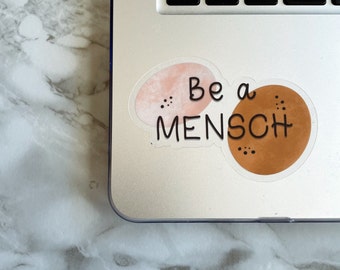 Könnte beinhalten: Ein Aufkleber mit zwei abstrakten Formen, einer rosa und einer braunen, mit dem Text "Be a Mensch" in Schwarz.