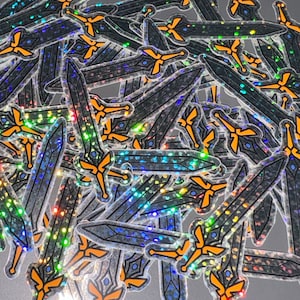 Op de afbeelding: Een stapel iriserende holografische stickers met een zwaardontwerp met een gouden handvat. De stickers zijn in een willekeurig patroon gerangschikt en zijn allemaal in dezelfde richting gericht.