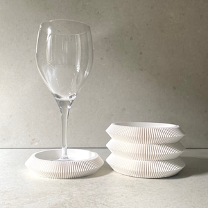 Puede incluir: Posavasos apilables blancos y geométricos con una copa de vino sobre un posavasos a juego. Los posavasos tienen un diseño acanalado.