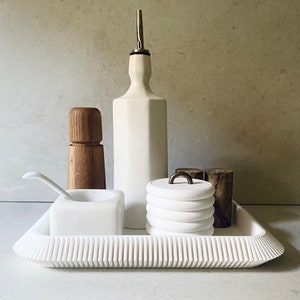 Könnte beinhalten: Weiße Keramik-Schale mit geriffeltem Rand, mit einer weißen Keramik-Öl-Flasche mit silbernem Ausgießer, einem weißen Keramik-Salz- und Pfefferstreuer-Set und einem weißen Keramik-Glas mit einem Holzdeckel.