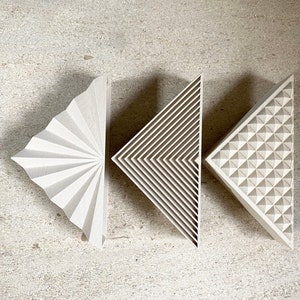 Puede incluir: Tres piezas de decoración de pared geométricas triangulares blancas con diferentes patrones. La primera pieza tiene un patrón de abanico, la segunda pieza tiene un patrón de rayas y la tercera pieza tiene un patrón de diamante.