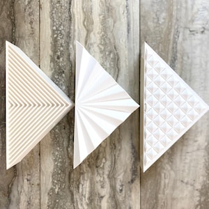 Puede incluir: Tres piezas de decoración de pared triangulares blancas con diferentes patrones en relieve. La primera pieza tiene un patrón lineal, la segunda pieza tiene un patrón de abanico y la tercera pieza tiene un patrón de diamante.