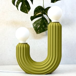 May include: A modern, olive green table lamp with a U-shaped design. The lamp features two white globe light bulbs and a ribbed texture. The lamp is connected to a white cord and sits on a white surface. A green plant with white spots is in the background.