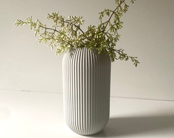 Può includere: Un vaso di ceramica bianco con nervature verticali, che contiene un mazzo di piccoli fiori bianchi con steli verdi.