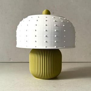 May include: A contemporary table lamp featuring a white, textured lampshade with small, raised dots. The base is a ribbed, cylindrical form in a yellow-green hue. A small, round, yellow-green detail is on top of the shade.