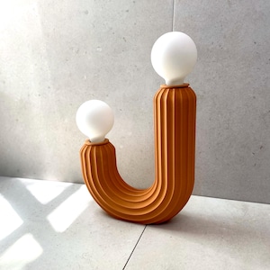 May include: A terracotta lamp shaped like the letter J with two white light bulbs. The lamp has a ribbed texture.