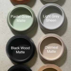 May include: Eight round, decorative containers with a textured, ridged edge. The containers are in various colors and finishes, including orange matte, white sheen, pastel green matte, light grey sheen, black wood matte, oatmeal matte, black sheen, and natural wood matte.