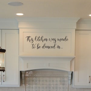 May include: A white kitchen with white cabinets and a white countertop. There are two black chandeliers hanging from the ceiling. The backsplash is white tile with a pattern. The text "This kitchen was made to be danced in..." is written on the wall above the stove.