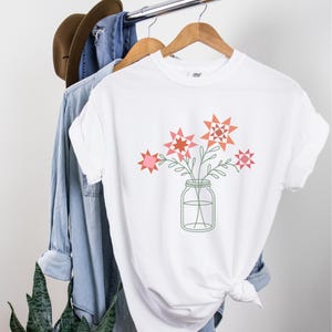 May include: White t-shirt featuring a graphic of a mason jar filled with pink and orange star-shaped flowers.