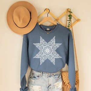 May include: A blue crewneck sweatshirt featuring a large white starburst design. The starburst has a detailed paisley pattern. A tan hat hangs on the wall above the sweatshirt.