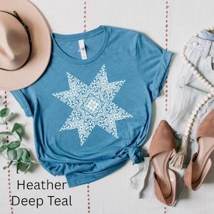 Puede incluir: Una camiseta de color azul teja jaspeado con un diseño de estrella blanca. La estrella tiene un patrón detallado e intrincado.