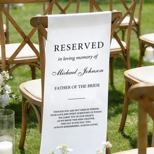 May include: A white banner with the word "RESERVED" and text honouring Michael Johnson, "FATHER OF THE BRIDE," hangs on a wooden chair. The banner includes a heartfelt message. The scene includes wooden chairs, white flowers, and candles.