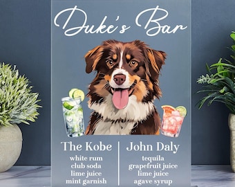 Op de afbeelding: Een helder acryl bord met de tekst "Duke's Bar" en een hondenportret. Het toont cocktailrecepten "The Kobe" en "John Daly" met ingrediënten. De achtergrond is een gedempt blauw.