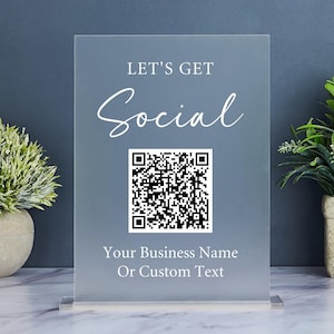 May include: A clear acrylic sign with the text "LET'S GET Social" and a QR code. Below the QR code is the text "Your Business Name Or Custom Text". The sign is displayed on a marble surface with small potted plants on either side.