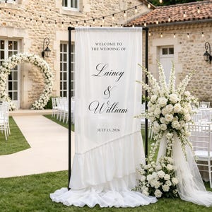 Può includere: Un cartello di benvenuto per matrimonio in tessuto bianco su una cornice in metallo nero con il testo "WELCOME TO THE WEDDING OF Lainey & William JULY 15, 2026" e un bordo inferiore arricciato, accanto a una grande composizione di rose bianche.