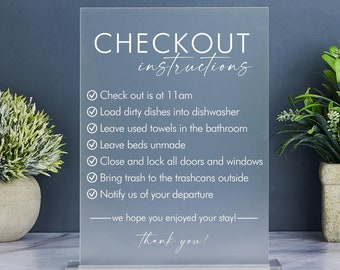 Puede incluir: Un cartel de acrílico blanco con el texto "CHECKOUT instructions" y una lista de instrucciones para los huéspedes que se registran en una propiedad de alquiler. Las instrucciones incluyen: Check out is at 11am, Load dirty dishes into the dishwasher, Leave used towels in the bathroom, Leave beds unmade, Close and lock all doors and windows, Bring trash to the bins outside, Notify us of your departure. El cartel también incluye el texto "we hope you enjoyed your stay!" y "Thank you!"