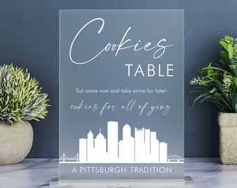 Può includere: Un cartello in acrilico trasparente con testo bianco che recita "Cookies Table" e "Mangiane subito e prendine qualcuna per dopo! Biscotti per tutti voi. Una tradizione di Pittsburgh." Il cartello presenta una silhouette bianca dello skyline di Pittsburgh.