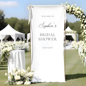 Puede incluir: Una pancarta blanca con el texto "Welcome to Sophia's Bridal Shower" y la fecha 10.05.2025. La pancarta forma parte de una ceremonia de boda o despedida de soltera, con arreglos florales y carpas blancas en el fondo.