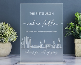 May include: A clear acrylic sign with a white outline of the Pittsburgh skyline and the text "THE PITTSBURGH cookie table Eat some now and take some for later! cookies for all of yinz".