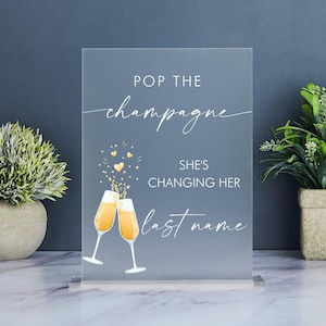Könnte beinhalten: Ein klares Acrylschild mit dem Text "Pop the champagne, she's changing her last name" in weißer Schrift. Zwei goldene Champagnergläser sind mit goldenem Konfetti und Herzen dargestellt.