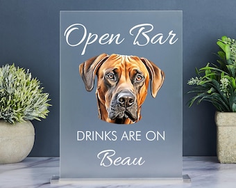 Op de afbeelding: Een helder acryl bord met de tekst "Open Bar Drinks Are On Beau" in witte letters. Een bruin en beige hondenhoofd bevindt zich in het midden van het bord.