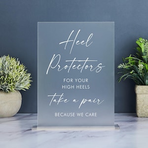 May include: A frosted acrylic sign with white text that reads "Heel Protectors for your high heels Take a pair because we care". The sign is on a white stand and is in front of a gray wall.
