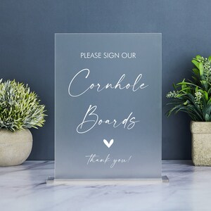 May include: A clear acrylic sign with white text that reads "Please sign our Cornhole Boards Thank you!" with a small white heart symbol.