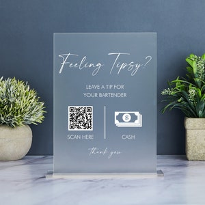 Puede incluir: Un letrero de acrílico esmerilado con el texto "Feeling Tipsy?" y "Leave a tip for your bartender." Incluye un código QR y un icono de efectivo. El letrero se muestra sobre una superficie de mármol con vegetación en el fondo.