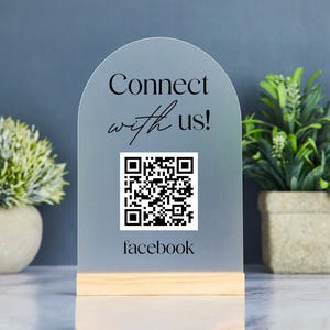 May include: A frosted acrylic sign with a wooden base. The sign has an arched top and features the text "Connect with us!" above a QR code and the word "facebook". Two small potted plants flank the sign.