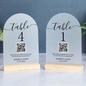 Peut inclure: Deux pancartes de numéro de table en acrylique transparent avec un sommet en arc arrondi. Les pancartes comportent un numéro de table noir, un code QR noir et blanc et le texte "Veuillez partager vos photos avec nous. Scannez simplement le code QR et téléchargez-les. SOPHIE & DAVID 12.24.2024". Les pancartes sont sur des bases en bois.