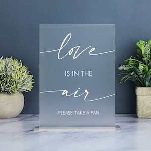May include: A frosted acrylic sign with the words "love is in the air" in white script. The sign also includes the text "PLEASE TAKE A FAN". The sign is displayed on a marble surface, with greenery in the background.