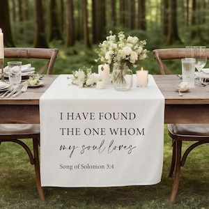 Op de afbeelding: Een witte tafelloper met de tekst "I HAVE FOUND THE ONE WHOM my soul loves" en "Song of Solomon 3:4". De loper ligt op een houten tafel die gedekt is voor een maaltijd, met bloemen en kaarsen.
