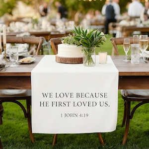 以下が含まれることがあります： 「WE LOVE BECAUSE HE FIRST LOVED US. 1 JOHN 4:19」のテキストが入った白いテーブルランナーが木製のテーブルに置かれています。ケーキ、緑、キャンドルがテーブルに配置され、結婚式やイベントにセットされています。