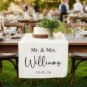May include: A white table runner with the text "Mr. & Mrs. Williams" and the date "10.05.26" is displayed on a wooden table. The table is set for a wedding reception, with a cake, flowers, and candles.