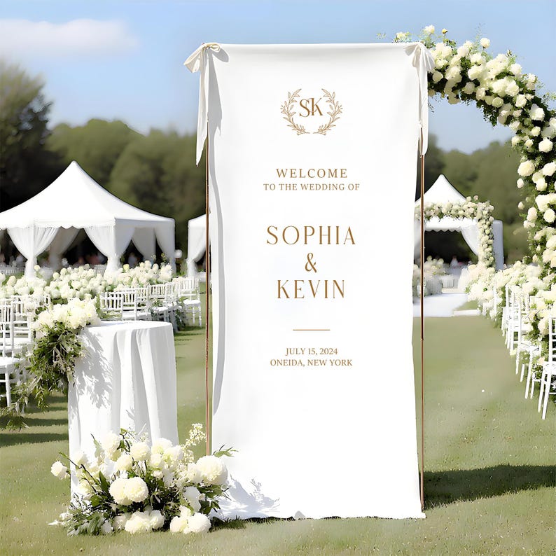 May include: A white wedding sign with gold lettering, welcoming guests to the wedding of Sophia & Kevin on July 15, 2024, in Oneida, New York. The sign features a floral wreath and is set up outdoors with a floral arch and white tents in the background.