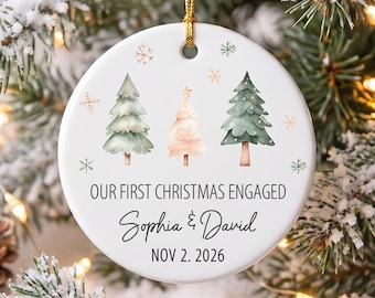 Puede incluir: Un adorno redondo de cerámica blanca con tres pinos estilo acuarela en verde y rosa suave, rodeados de pequeños copos de nieve. El texto dice: OUR FIRST CHRISTMAS ENGAGED, Sophia & David, y la fecha NOV 2. 2026, con un cordón dorado para colgar.