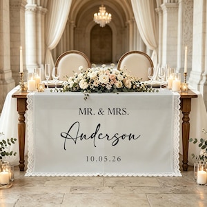 May include: A white fabric table runner with lace trim featuring the black printed text MR. & MRS. Anderson and the date 10.05.26. The runner is draped over a wooden table decorated with a floral centerpiece, candles, and glassware in a formal setting.