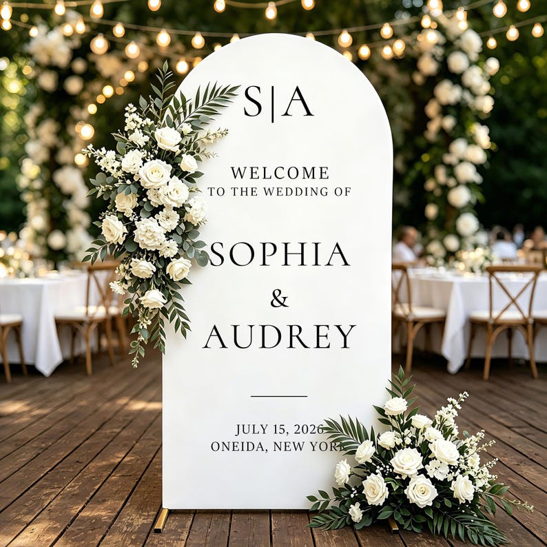 Puede incluir: Un letrero de bienvenida de boda en forma de arco blanco con el texto negro S|A, WELCOME TO THE WEDDING OF, SOPHIA & AUDREY, JULY 15, 2026, ONEIDA, NEW YORK. El letrero est&aacute; decorado con rosas blancas y follaje verde en la parte superior izquierda e inferior derecha.