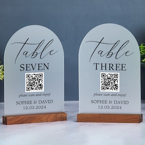 May include: Two frosted acrylic table signs with arched tops and wooden bases. Each sign displays "Table" in script, a table number, a QR code, and the text "please scan and enjoy!" along with a date and names.