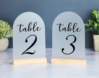 May include: Two frosted acrylic table number signs with wooden bases. The signs are white with black text that reads "Table 2" and "Table 3".