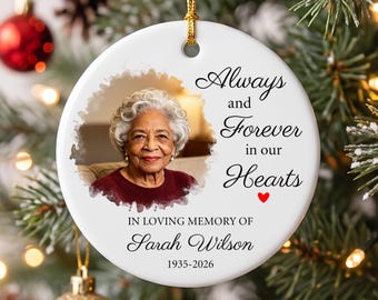 May include: A round white ceramic memorial ornament featuring a photograph of an elderly person with gray hair wearing a maroon sweater. The text reads Always and Forever in our Hearts, IN LOVING MEMORY OF, Sarah Wilson, and 1935-2026, with a small red heart.