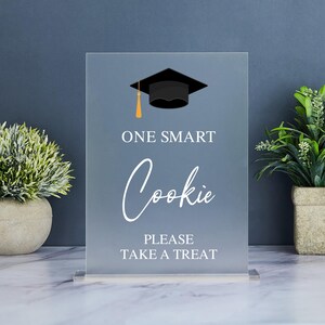 May include: A clear acrylic sign with a black graduation cap graphic and the text "One Smart Cookie Please Take A Treat".